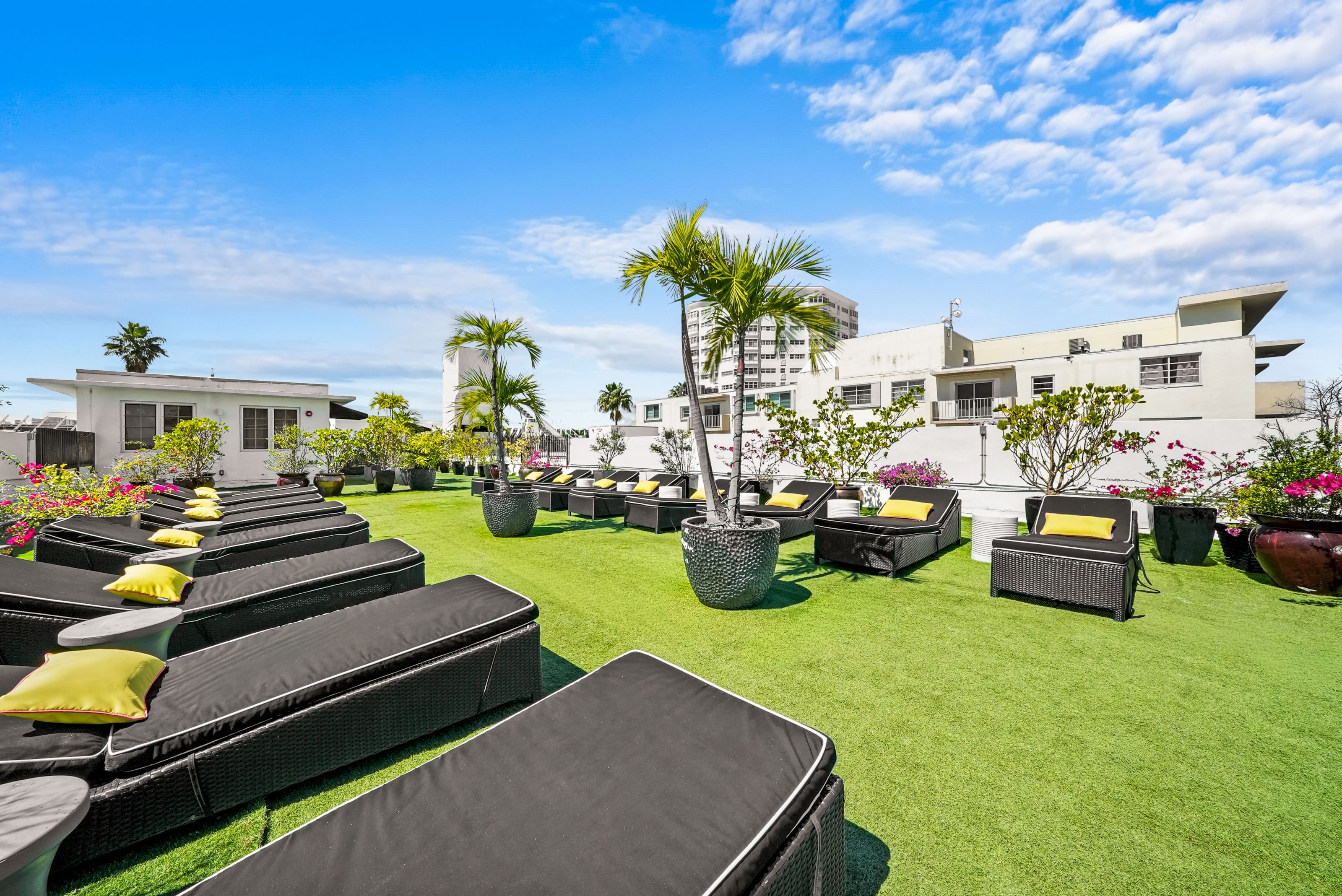 A lounge area with black wicker chairs and yellow pillows on artificial grass, surrounded by potted plants and a white building in the background.