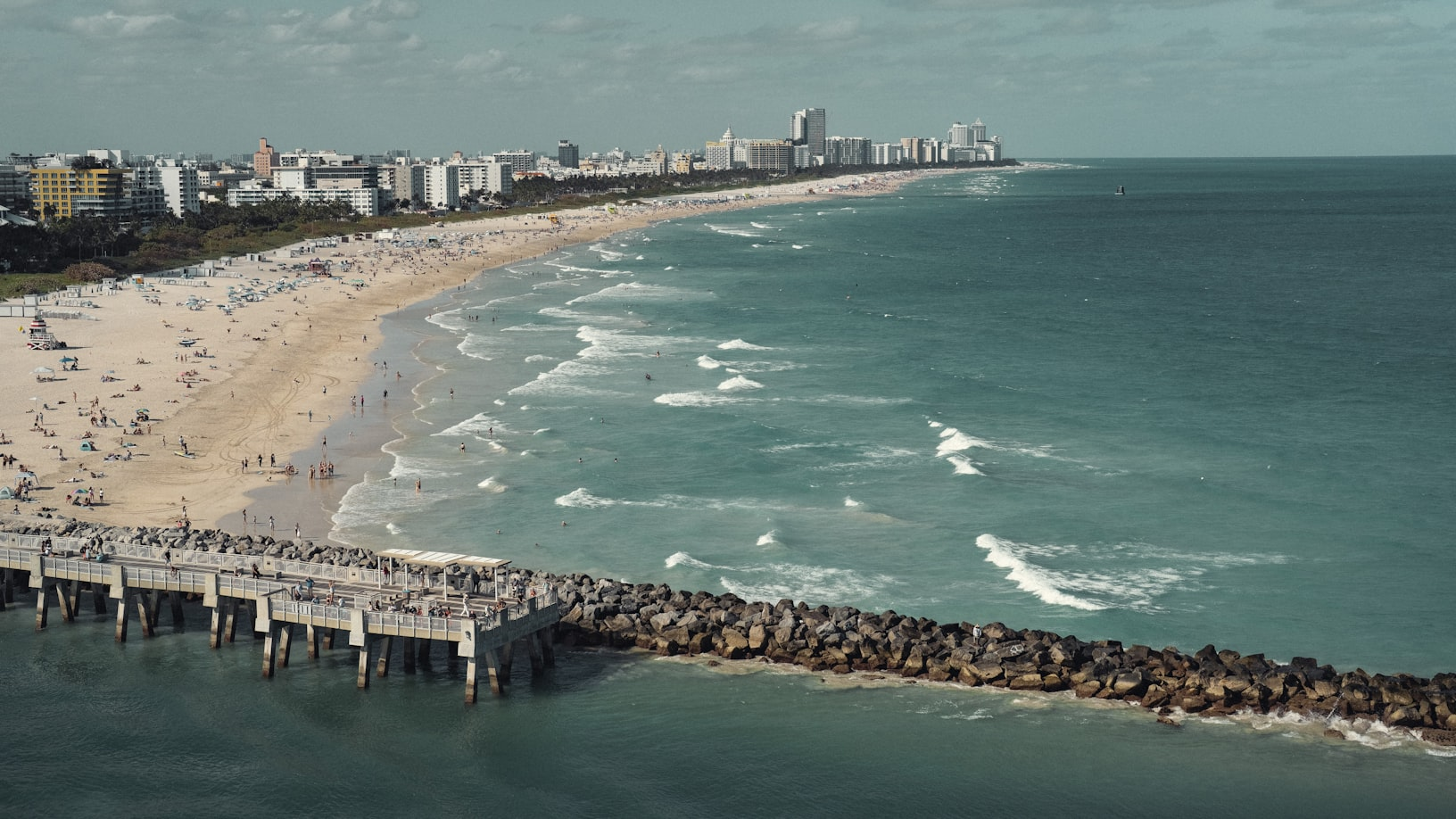 A wide-angle view of a beach with a pier, showing the ocean, sandy beach, and city skyline in the distance.