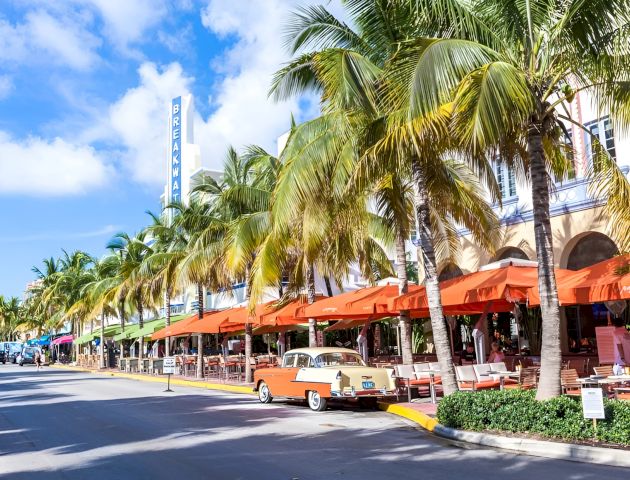 A vintage car parked in front of a restaurant patio with orange umbrellas and palm trees.