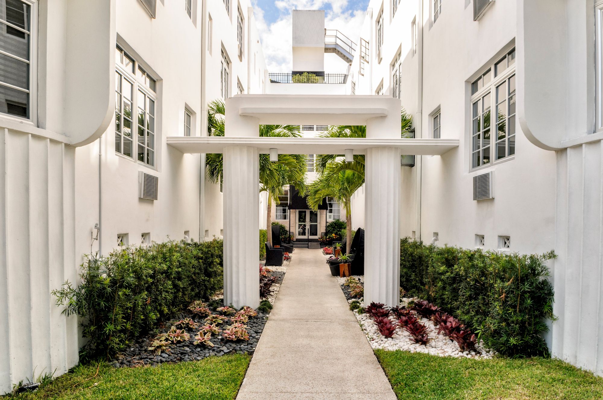 A bright courtyard walkway with white columns, trimmed greenery, and flowering plants between tall apartment buildings.