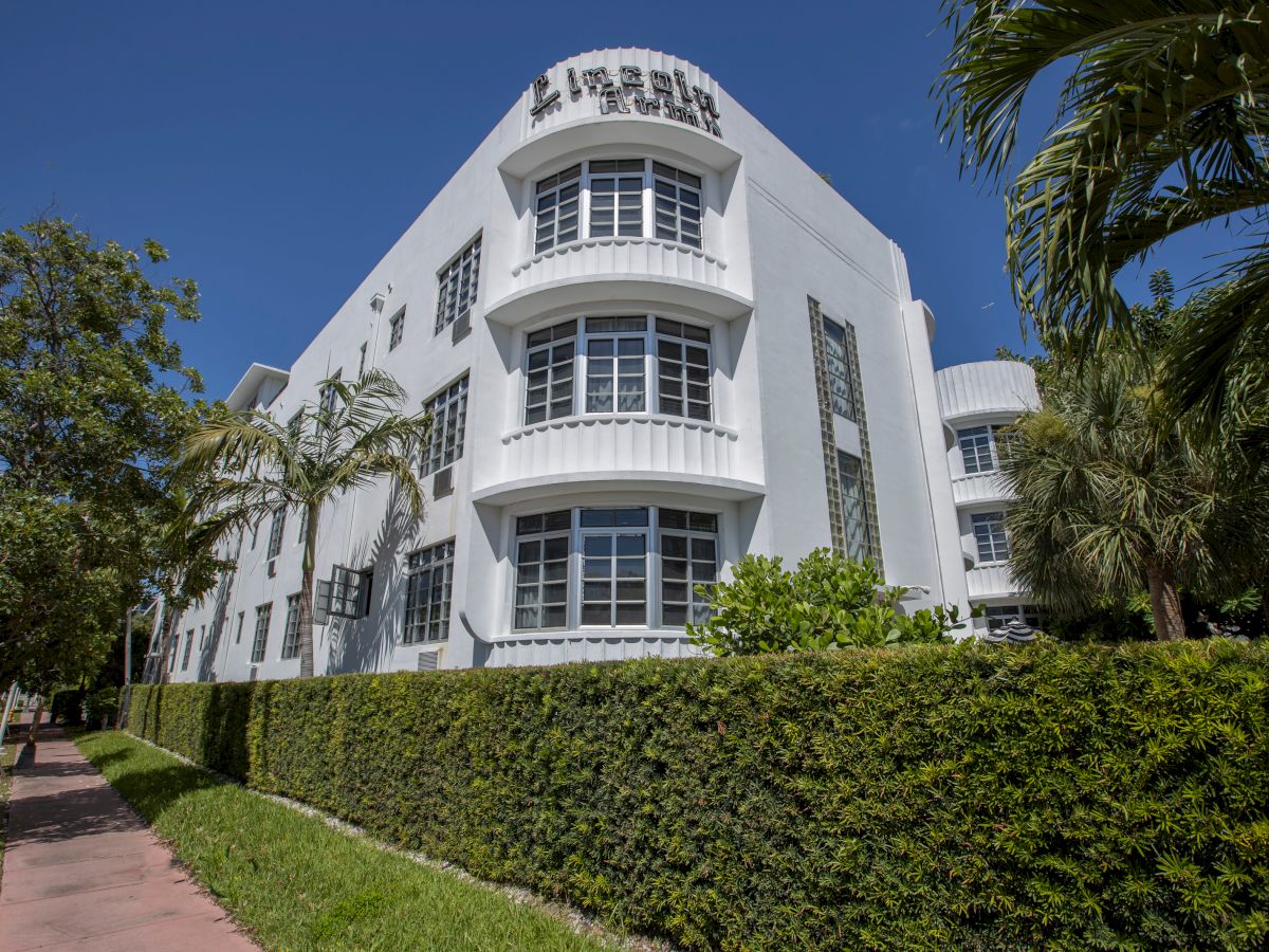 A modern white multi-story hotel building with a rooftop sign and bay windows, surrounded by palm trees and trimmed hedges.