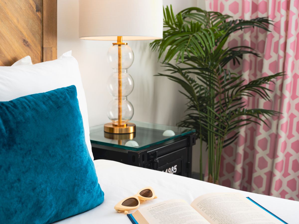 A bright bedroom corner with a blue pillow, open book, tall gold lamp, potted palm, and patterned curtains.