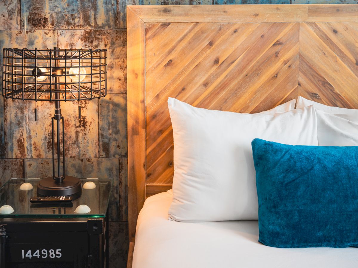 A modern hotel room scene with a bed, white and blue pillows, a wooden headboard, and a warm table lamp against a metal wall.