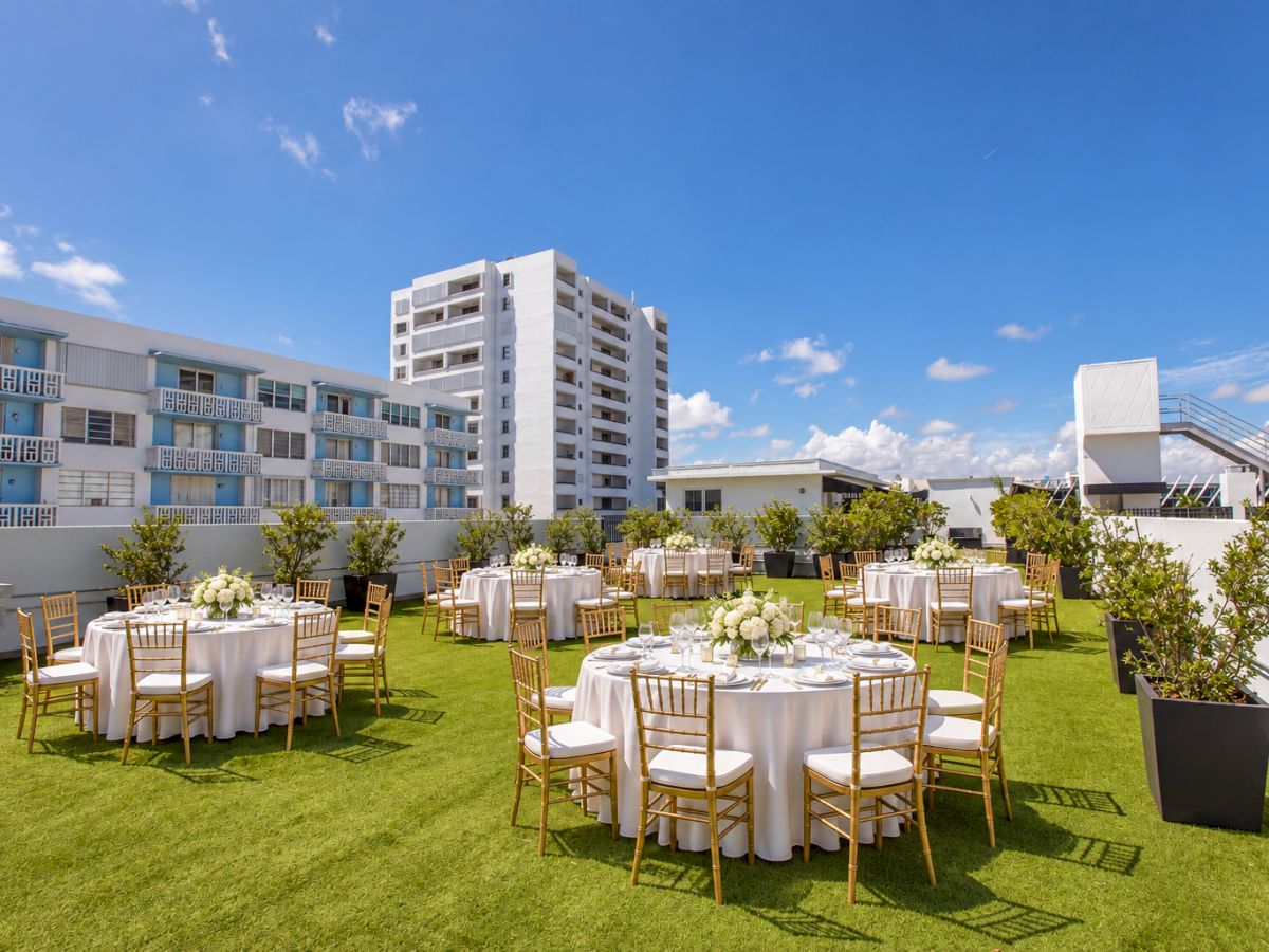 A hotel courtyard wedding setup with white tablecloths, gold chairs, flower centerpieces, and apartment buildings under a blue sky.
